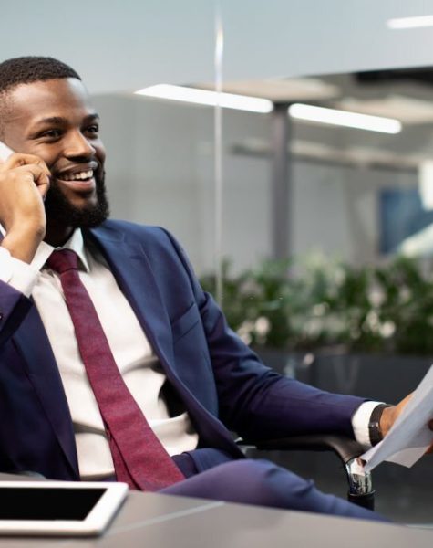 Happy african american businessman sitting at workdesk with modern digital tablet with blank screen on at office, having phone call with business partner, holding papers, panorama with copy space